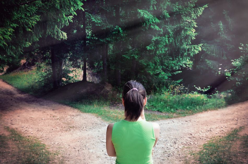 Eine Frau steht im Wald an einer Weggabelung und blickt auf die zwei Wege